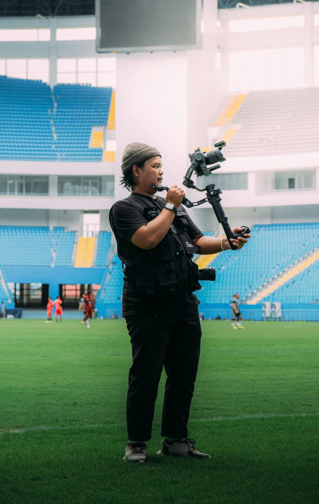 Tournaments A videographer holds a camera stabilizer on a sports field in an empty stadium.